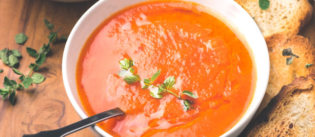 Tomato soup in a white bowl with bread and herbs on a wooden surface